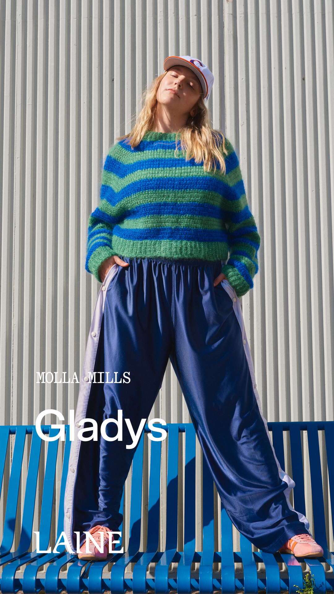Person wearing a green and blue striped Gladys sweater designed by Molla Mills and blue pants standing on a blue bench.