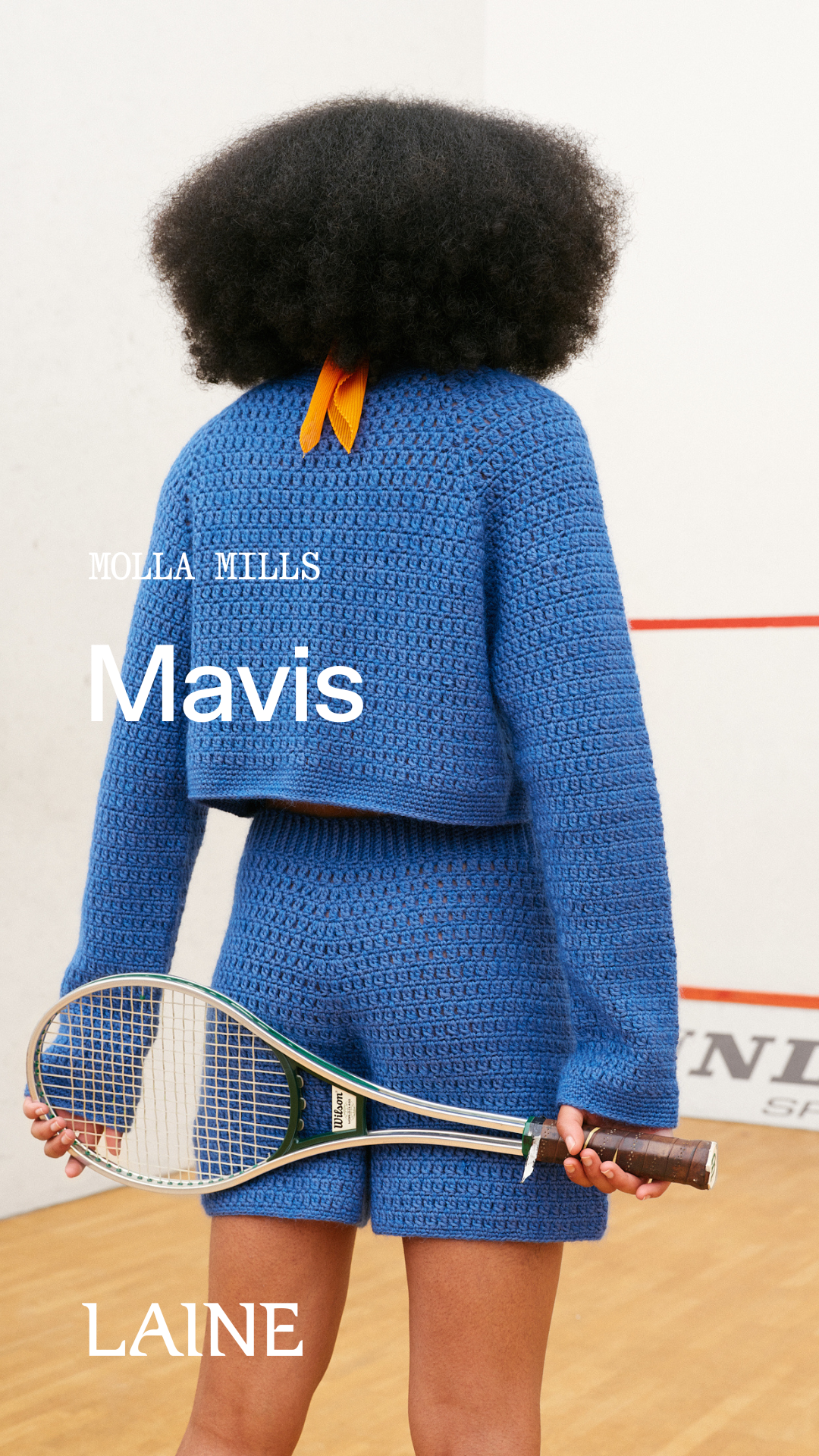 Person wearing a blue crochet Mavis  sweater and short set designed by Molla Mills holding a tennis racket on a wooden floor.