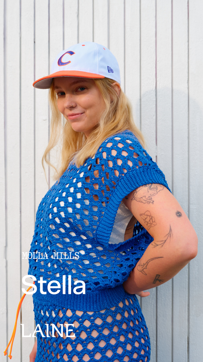 Woman wearing a blue crochet Stella  top and skirt set designed by Molla Mills and a cap against a white background