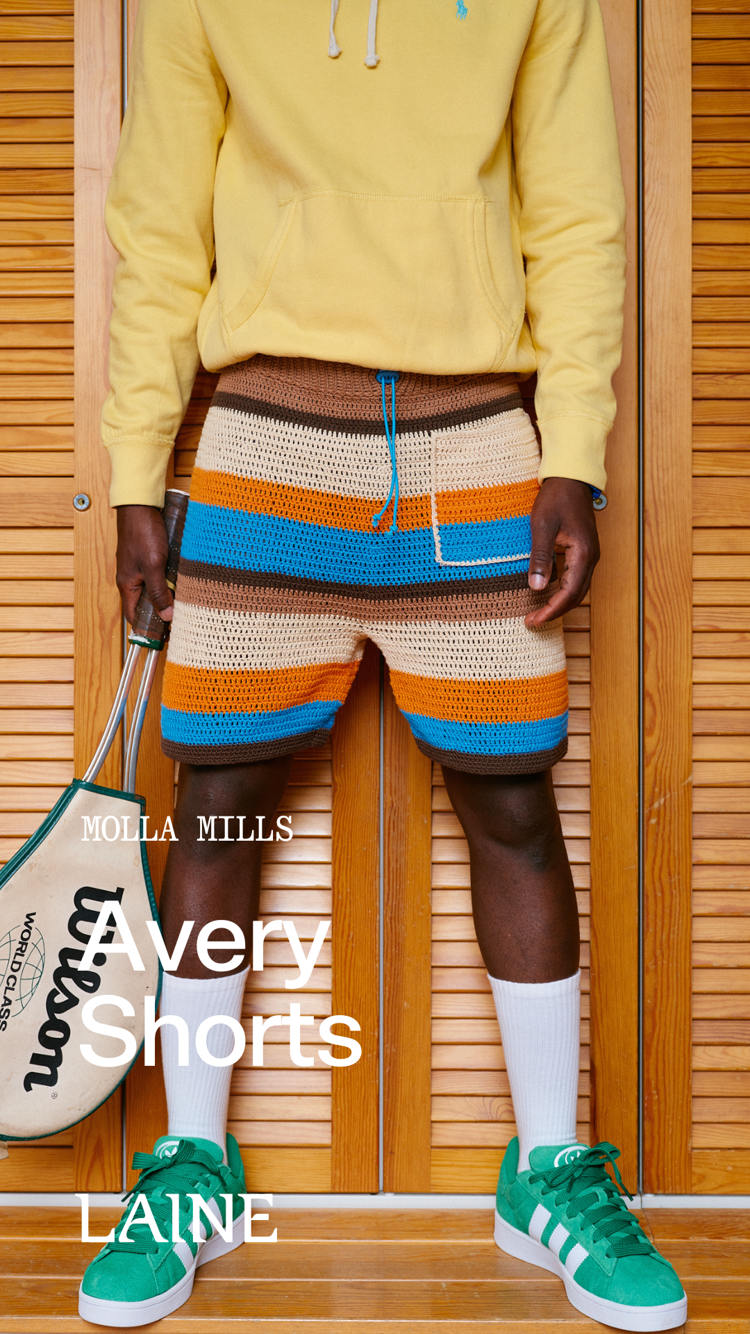 Person wearing a yellow hoodie, striped Avery shorts designed by Molla Mills, and green sneakers with a tennis racket, standing against a wooden panel background.