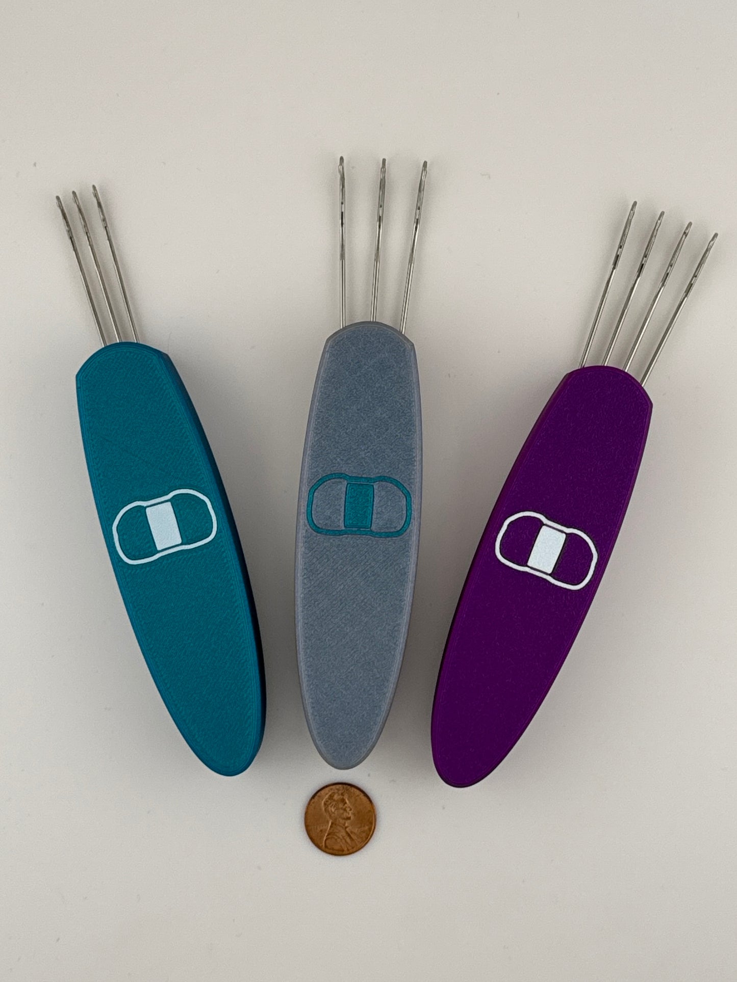 Three i-cord knitting tools in different colors (blue, gray, and purple) with stainless steel hooks, displayed with a coin for scale.