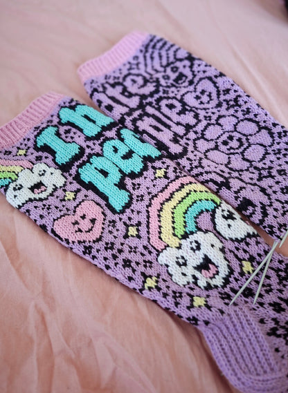 Knitted socks with text and patterns on a pink fabric background. One sock has colorful duplicate stitching the second does not
