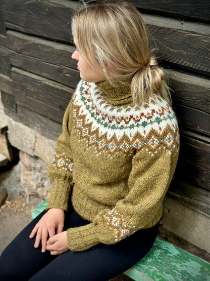 A woman wearing a hand-knit turtleneck sweater with a stranded colorwork pattern in olive green, cream, brown, and teal, sitting against a dark wooden wall.