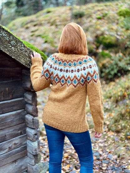 A woman viewed from behind wearing a yellow knit sweater with a colorful stranded colorwork circular yoke pattern in blue, brown, and cream, standing outdoors next to a rustic wooden cabin.