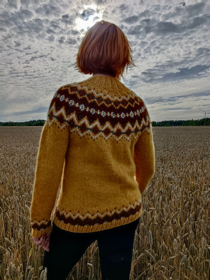 A person standing in a wheat field, viewed from behind, wearing a mustard yellow knit sweater with a brown and cream Nordic-style pattern across the shoulders and upper back, paired with black pants.