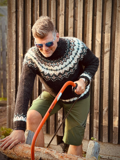 A man wearing a dark gray and cream stranded colorwork patterned knit sweater, olive green shorts, and sunglasses is seen sawing a log outdoors against a wooden fence.
