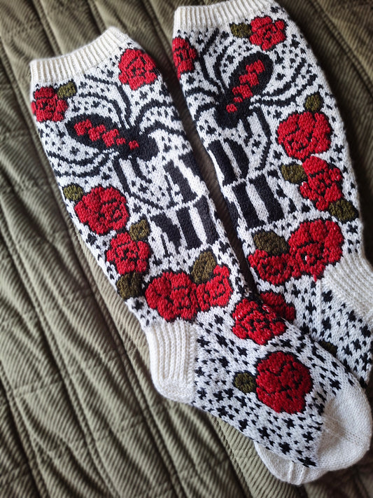 A pair of hand-knit socks featuring a detailed pattern of red roses, black and white stripes, and a butterfly, laid flat on a textured olive green blanket.