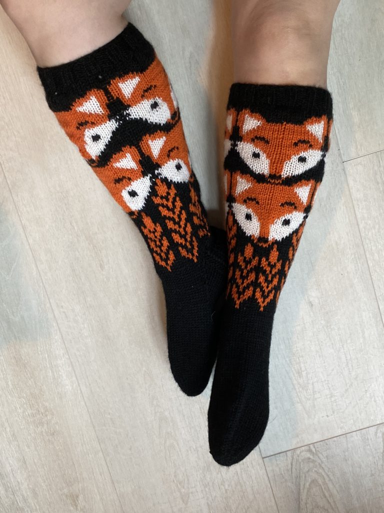 Below the knee knitted wool socks with a black background, orange fox faces, and wheat patterns